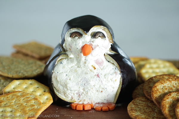 Image of cheeseball appetizer decorated like a penguin, sitting on a wood board with crackers