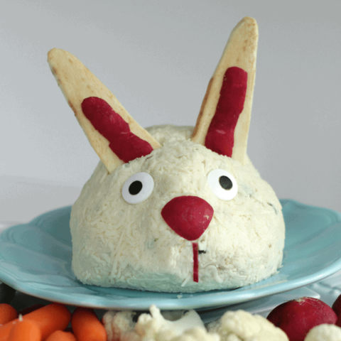 Cheeseball decorated to look like an Easter bunny sitting on a blue plate with vegetables around it