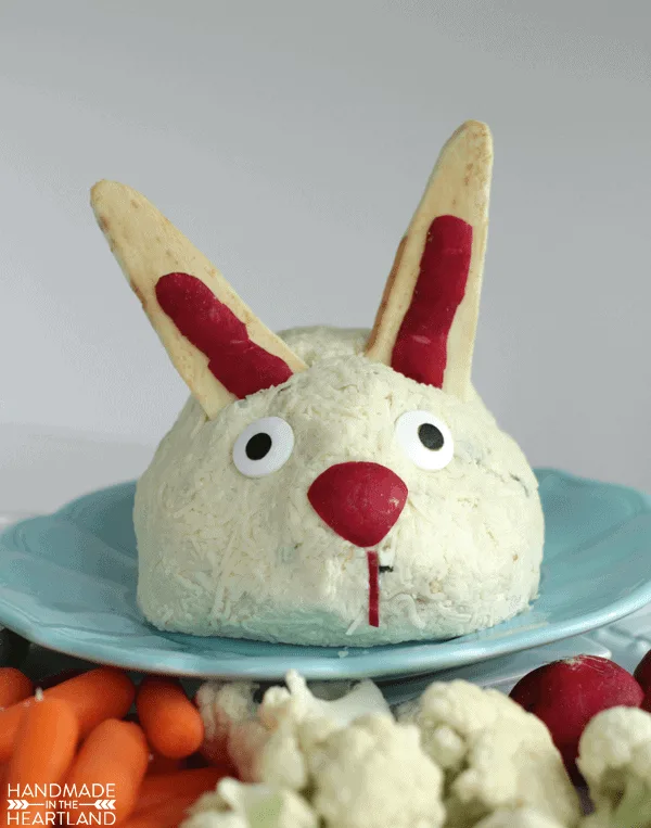 Cheeseball decorated to look like an Easter bunny sitting on a blue plate with vegetables around it