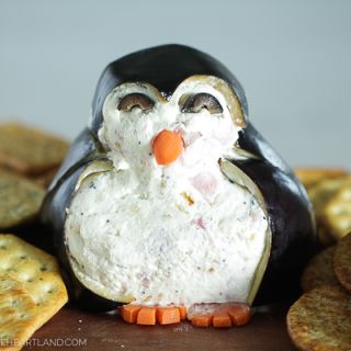 Image of cheeseball appetizer decorated like a penguin, sitting on a wood board with crackers