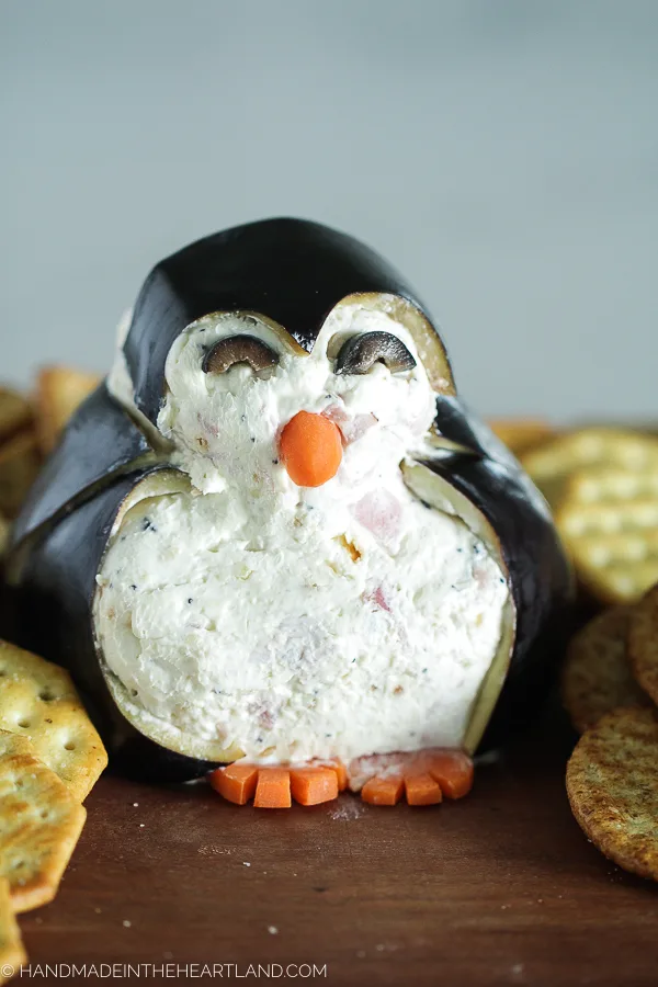 Cheese ball appetizer decorated to look like a penguin sitting on wood board with crackers