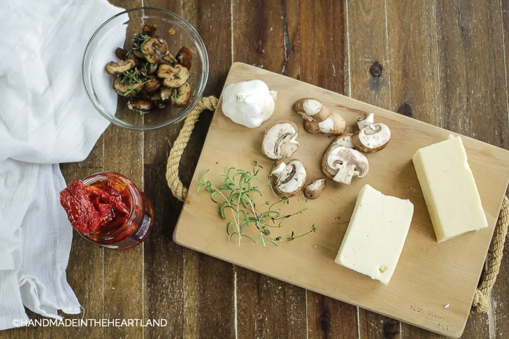 toppings for pizza with mushrooms, cheese, rosemary, sundried tomatoes sitting on wood board and wood table