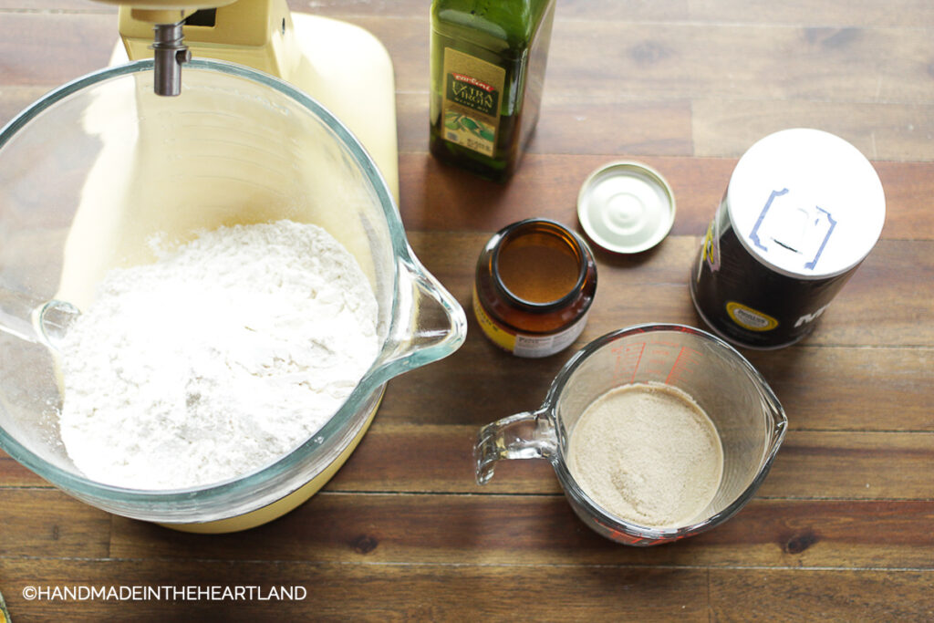 kitchen aid mixer with flour in the bowl with olive oil, yeast, salt and measuring cups sitting next to mixer